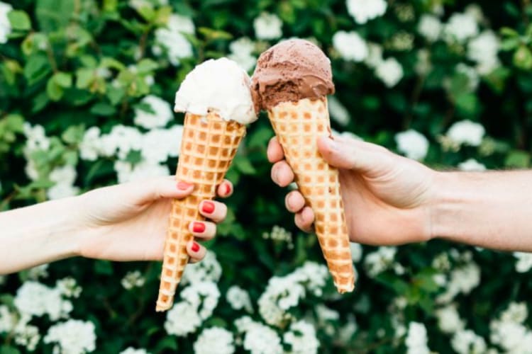 Two people holding ice cream cones next to a green hedge with white flowers