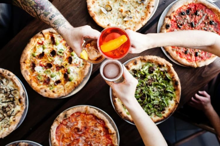 People cheersing drinks over a table with pizzas on it