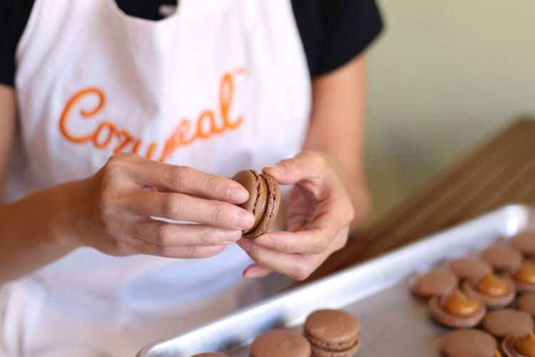 A person's hands holding a macaron, above a tray of macarons, during a cooking class