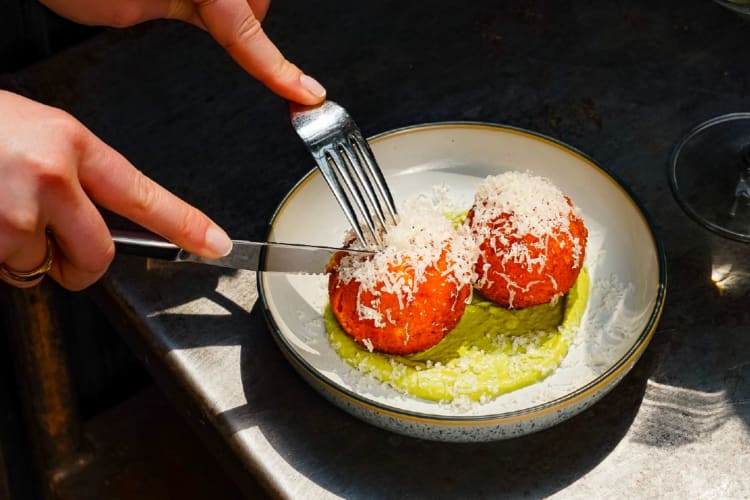 Hands cutting into two arancini balls topped with cheese with a knife and fork