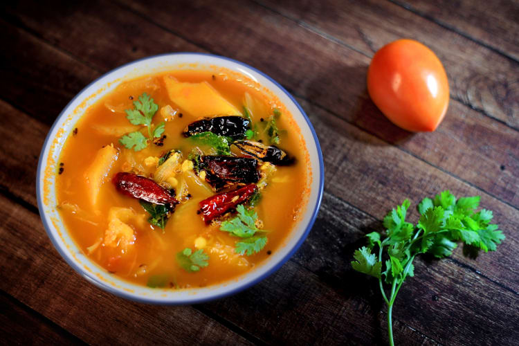 A South Indian food called Sambar in a bowl, next to a tomato and a herb