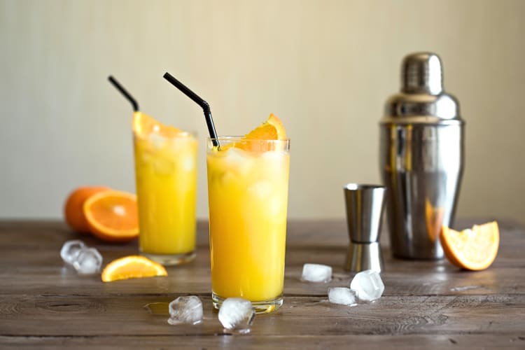 Two screwdriver vodka cocktails on a wooden surface with ice, orange slices, a shot pourer and a shaker