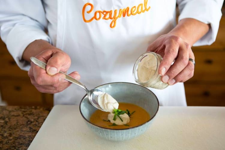 A chef in a Cozymeal apron putting cream on a bowl of soup