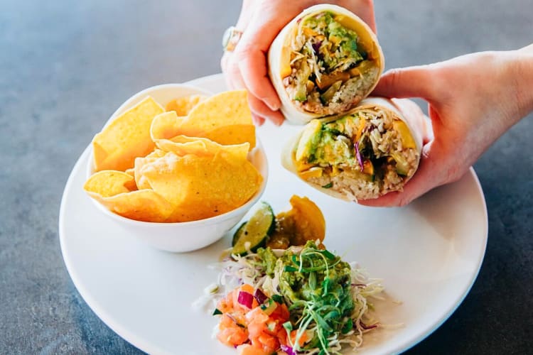 Person's hands holding two halves of a burrito next to chips and a salad
