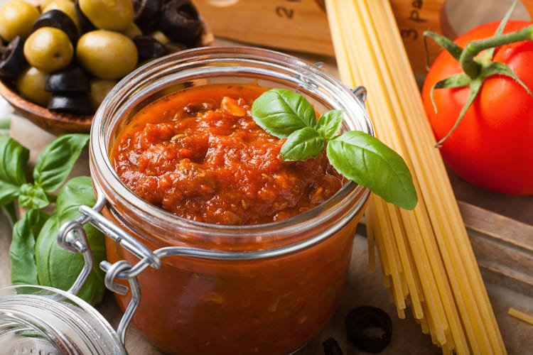 Glass jar with tomato pasta sauce, surrounded by pasta, tomatoes, olives and basil