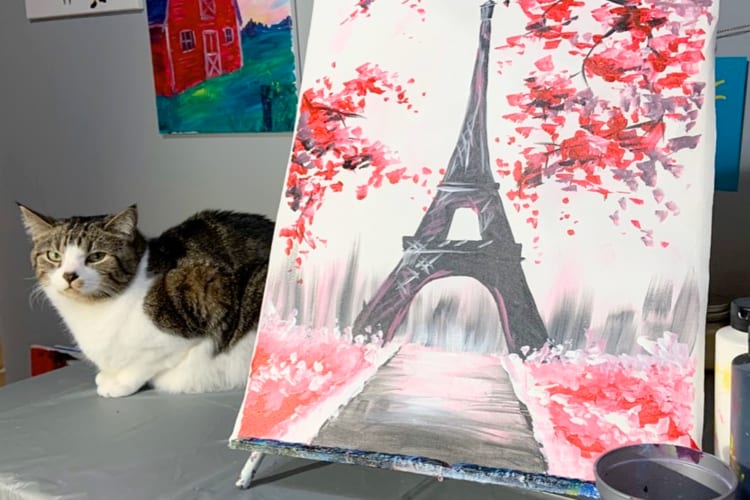 A white and tabby cat sitting next to an Eiffel Tower painting