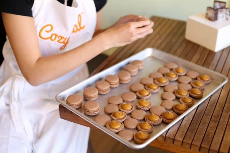 A person in a Cozymeal apron filling chocolate macaron shells with caramel