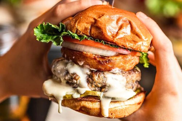 Person's hand holding a burger with pickles, tomato, lettuce, onion and an onion ring