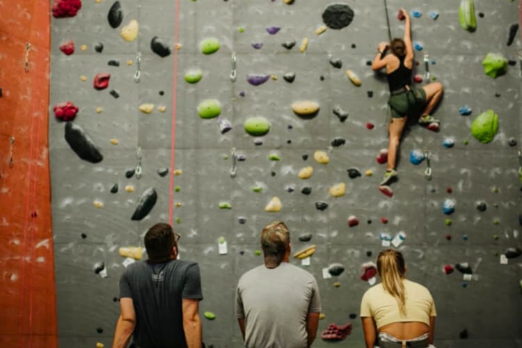 Three people watching another person climb an indoor rock wall