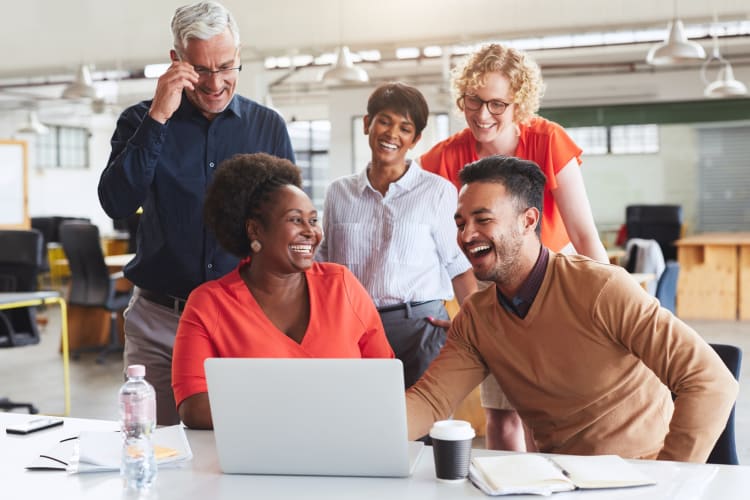 A group of co-workers laughing while sitting and standing around a desk