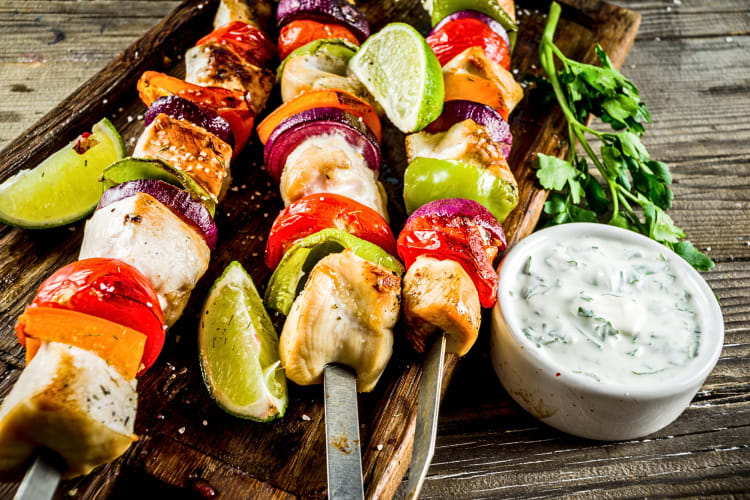 Three kebabs with lemon wedges on a wooden serving board, next to a bowl of sauce