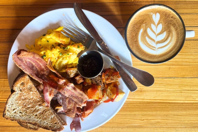 Breakfast with toast, bacon, potatoes and eggs next to a cappuccino 