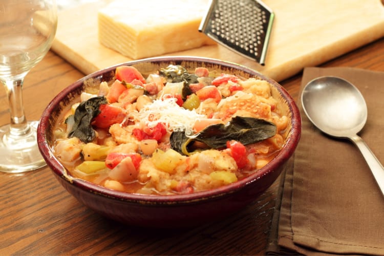 A bowl of bean and vegetable soup next to wine glasses and Parmesan