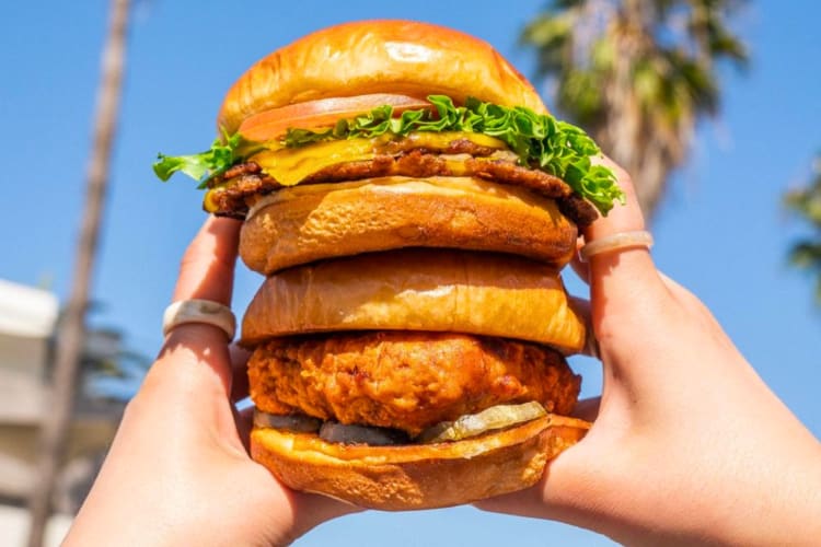 A person holding a veggie burger with palm trees in the background