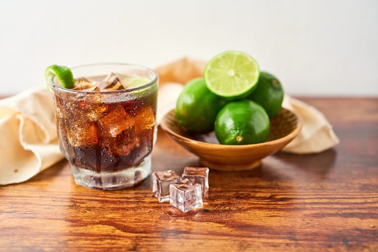 Vodka and cola, ice cubes and a bowl of limes, on a wooden surface