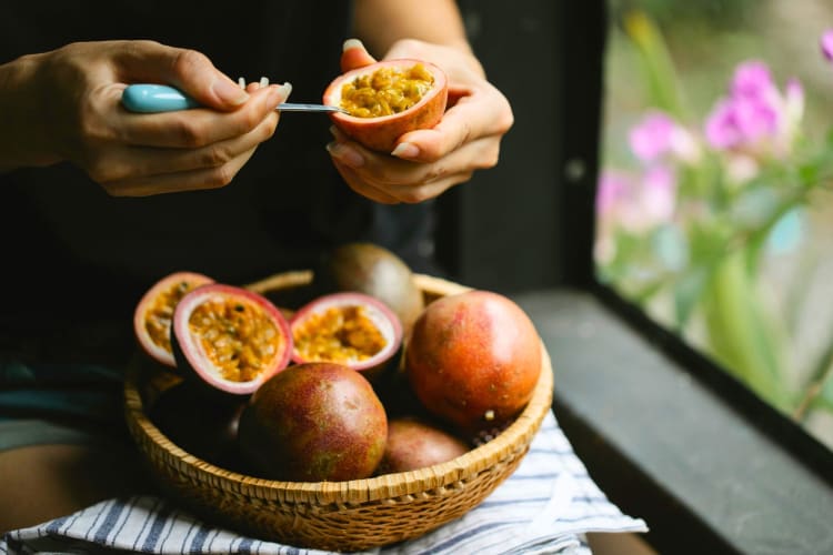 A person scooping the pulp out of a passion fruit next to a bowl of passion fruit