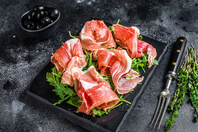 Prosciutto meat on top of a bed of leaves on a black serving board