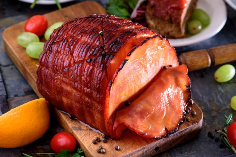 A cooked ham on a wooden board surrounded by pieces of fruit. 