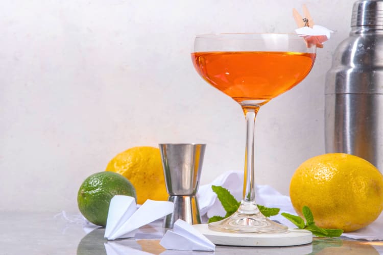 An orange cocktail next to lemons and limes and a small paper plane