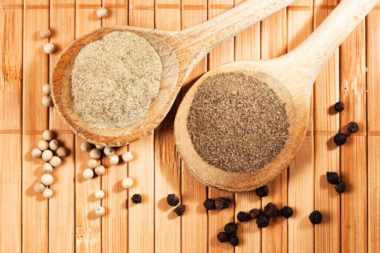 Ground white pepper and black pepper on wooden spoons surrounded by whole peppercorns