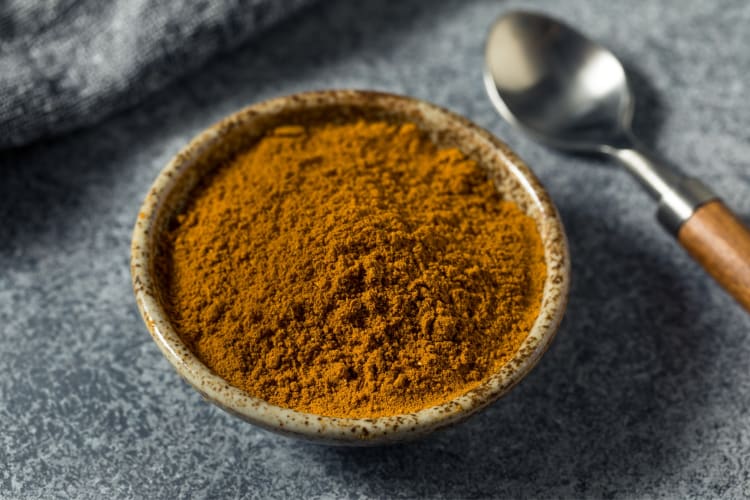 A bowl of cumin with a spoon next to it on a grey surface