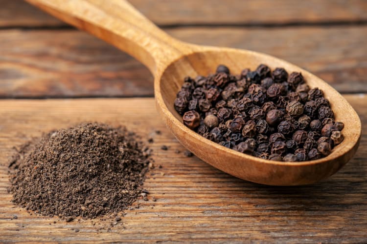 Whole black peppercorns on a wooden spoon next to ground black pepper on a wooden table