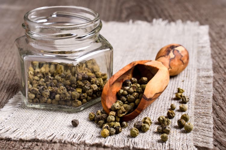 Green peppercorns in a glass jar next to green peppercorns on a wooden spoon