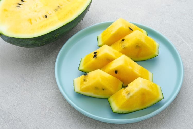 Triangles of cut yellow watermelon on a blue plate next to half a yellow watermelon