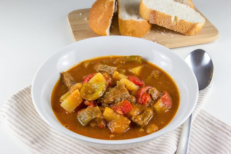 A bowl of potato goulash next to a spoon, in front of a wooden board with bread slices