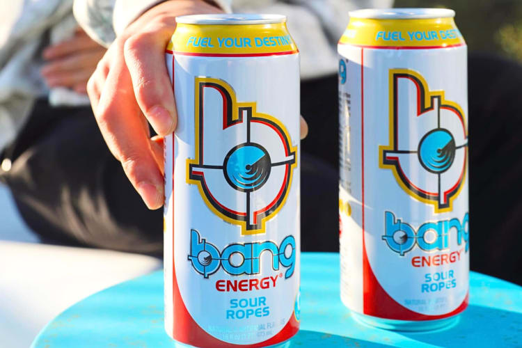 A hand holding a can of Bang Energy, next to another of the same can
