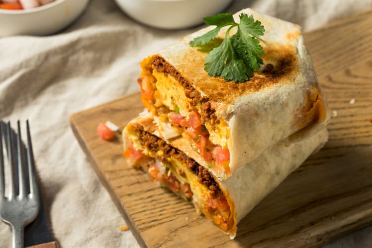 A breakfast burrito with meat, eggs and salsa on a board