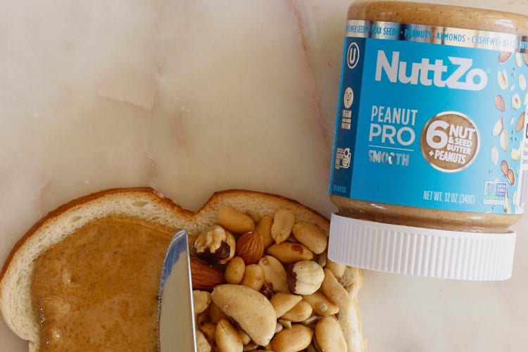 A bottle of Nuttzo Peanut Trail Mix Nut & Seed Butter next to a bread slice with nuts