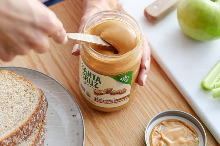 A hand scooping peanut butter with a knife from a Santa Cruz Organic bottle