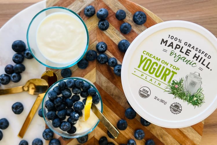 A tub of Maple Hill Creamery yogurt next to two glasses of yogurt and blueberries