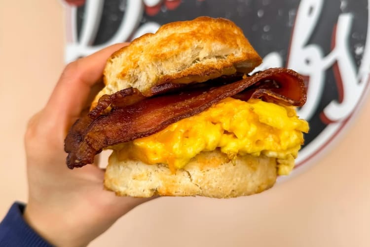 a hand holding up a breakfast biscuit sandwich with egg, cheese and bacon