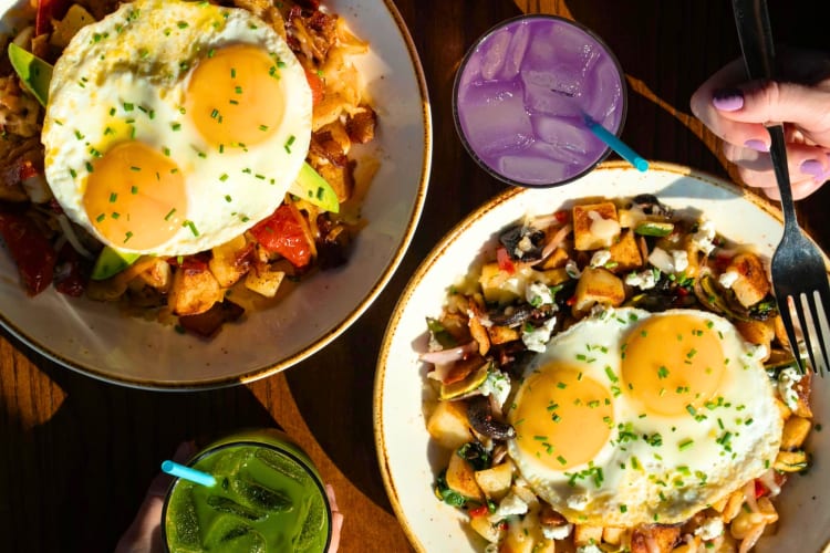 Two breakfast hash bowls on a table with green and purple drinks