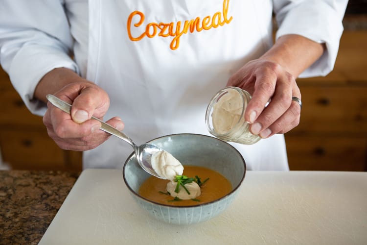 A chef, wearing a Cozymeal apron, putting cream on a soup