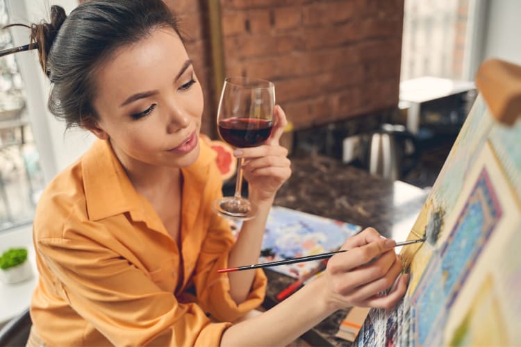 a woman painting on a canvas while holding a glass of red wine