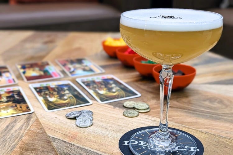 A cocktail with egg white foam next to playing cards and coins