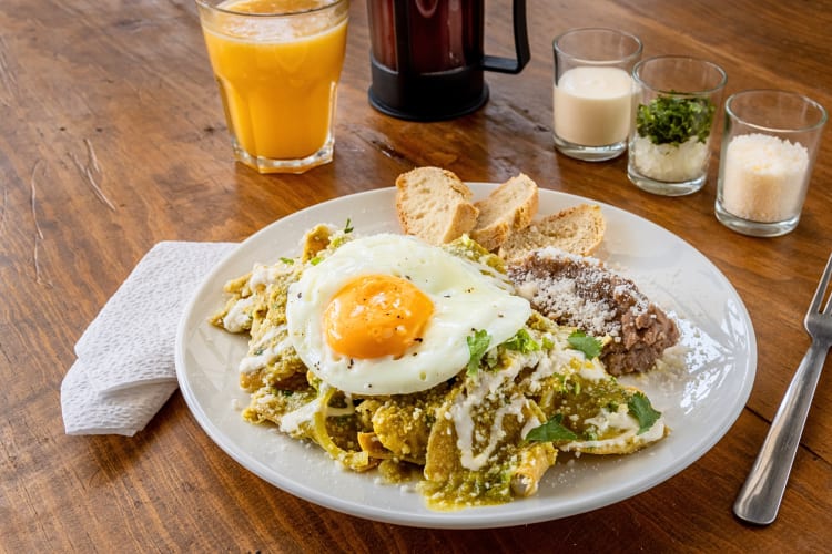 Eggs, chilaquiles and bread on a plate in front of a glass of orange juice
