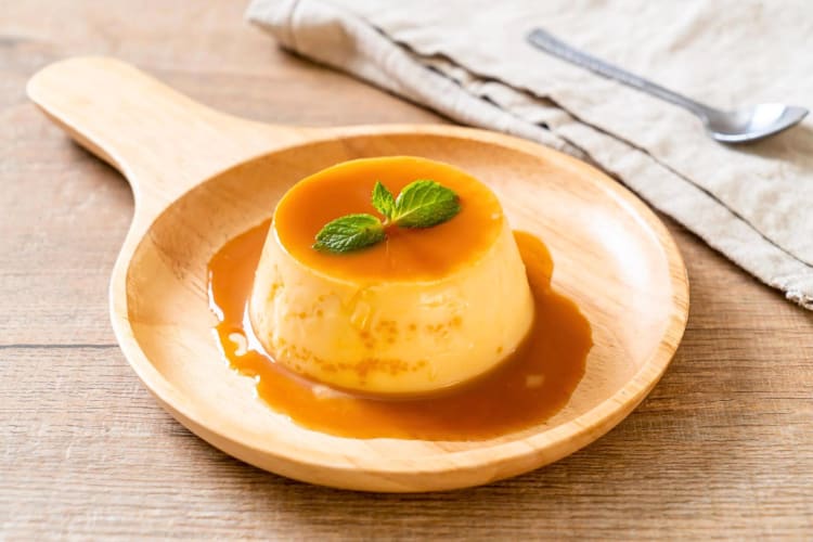 A portion of leche flan on a wooden plate, next to a spoon