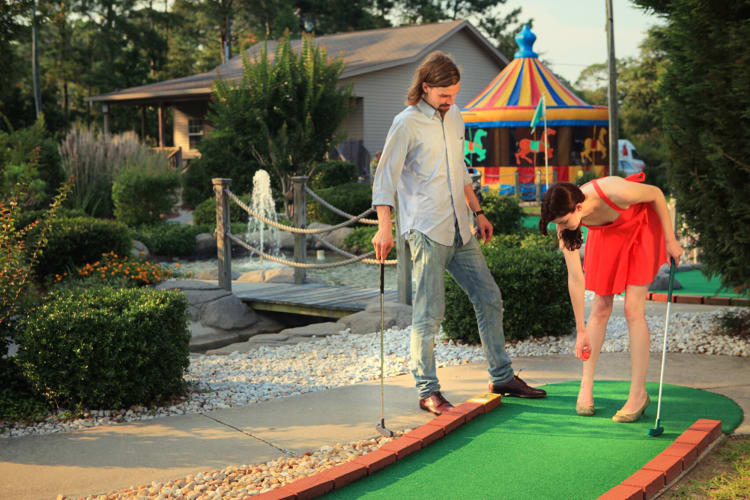 A man watching a woman place a golf ball during a mini golf game