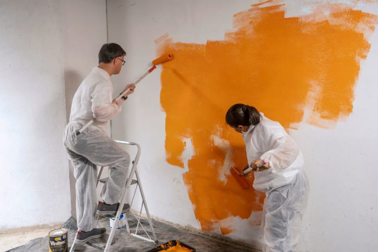 two people paint a wall in a house orange with rollers