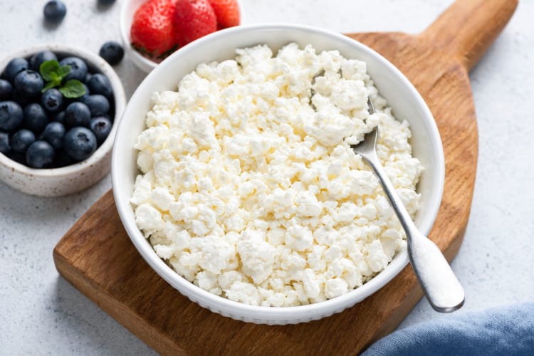 Cottage cheese crumbles in a white bowl with fruit in the background.