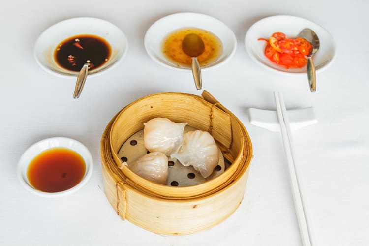 Three har gow shrimp dumplings in a bamboo basket, surrounded by sauces