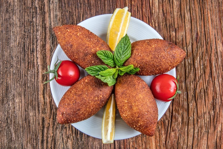 Aerial view of four İnegöl köfte on a plate with tomatoes, herbs and lemon wedges