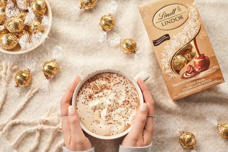 A box of Fudge Swirl Lindor chocolate flavor truffles next to a hand holding a mug