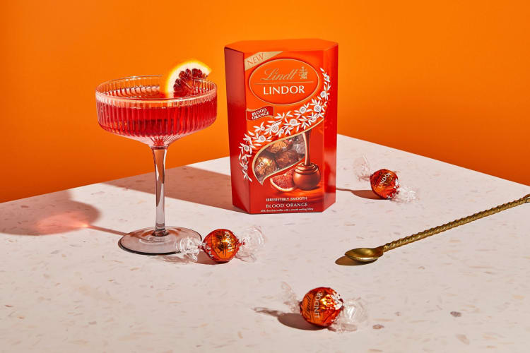 A box of Lindor Blood Orange truffles, a spoon and an orange drink in front of an orange backdrop