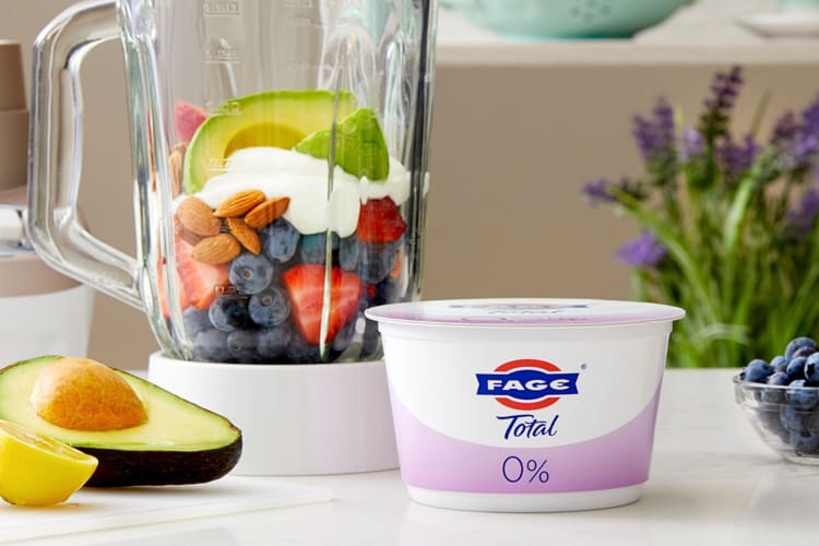 A tub of FAGE Total 0% low-calorie yogurt next to a blender with smoothie ingredients in it