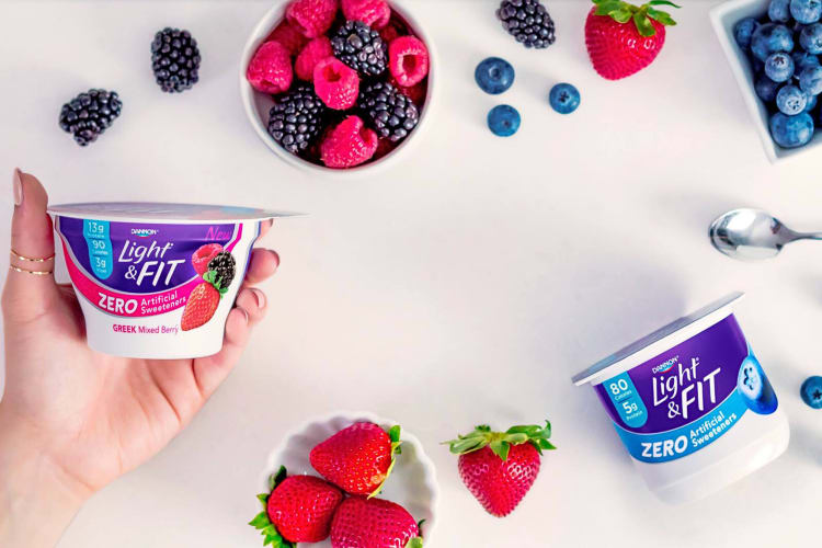 A hand holding a Dannon low-calorie yogurt, next to another one, surrounded by fruit